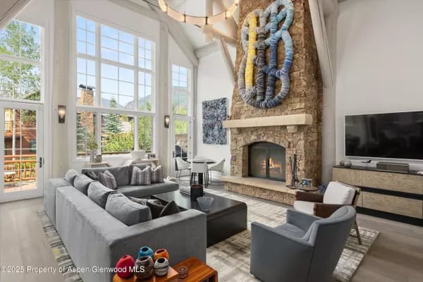 Living room with tall windows, gray seating, and a stone fireplace with sculptural wall art.