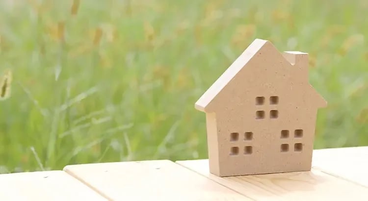 A small wooden house model set on a table with a grassy background. The scene evokes a sense of simplicity and nature.