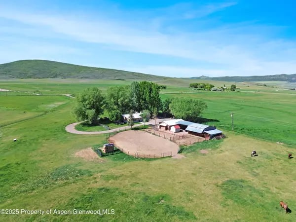 Aerial view of a rural property with farm buildings, a fenced arena, green pasture, and horses.