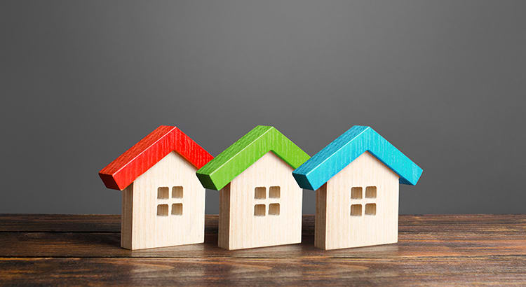 A conceptual image showing three small wooden houses with red, green, and blue roofs, placed on a wooden surface against a gray background. The image symbolizes real estate, housing, or home ownership.