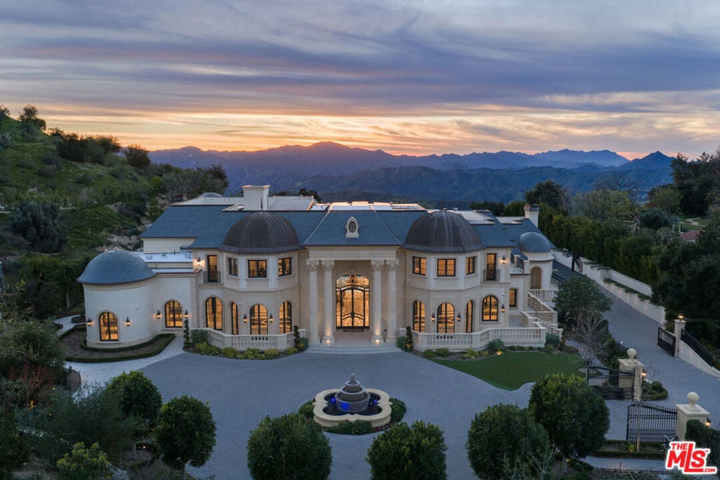 Luxury estate with twin domed wings and a circular fountain, set against mountains at sunset.