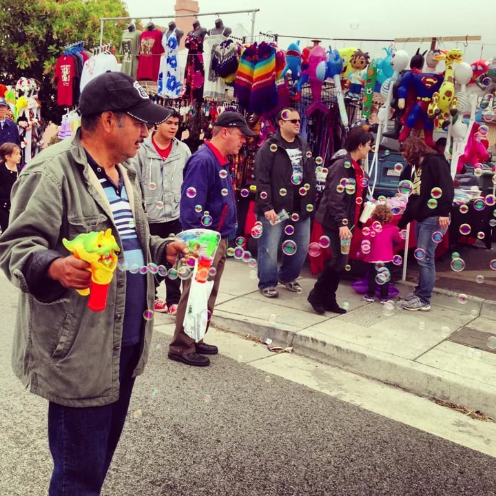 A man blowing bubbles with a novelty dinosaur bubble gun at an outdoor street festival with a line of people and a stall selling colorful merchandise and stuffed toys in the background.