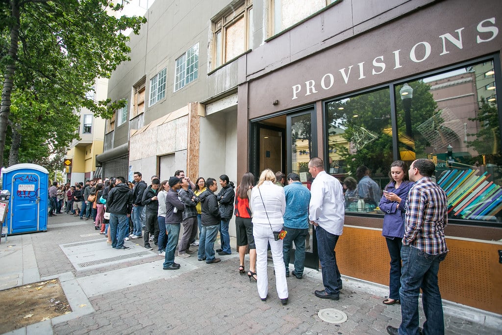 The exterior of State Bird Provisions at 1529 Fillmore Street in San Francisco, featuring a modern facade with large windows and the restaurant's name displayed above the entrance. The sidewalk is lined with planters, and the warm interior lighting creates an inviting atmosphere for passersby.