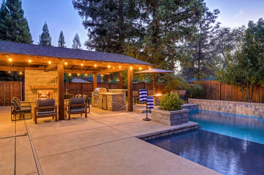 Backyard pool beside a covered patio with an outdoor kitchen and warm string lights at dusk.