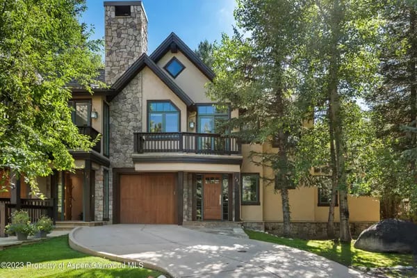 Two-story home with a stone chimney, upper balcony, and curved driveway, framed by mature trees.