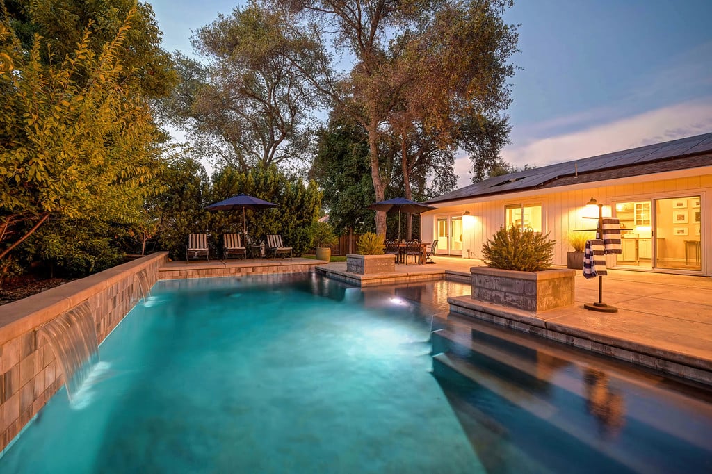 Backyard pool beside a single-story home, with patio seating and solar panels at dusk.