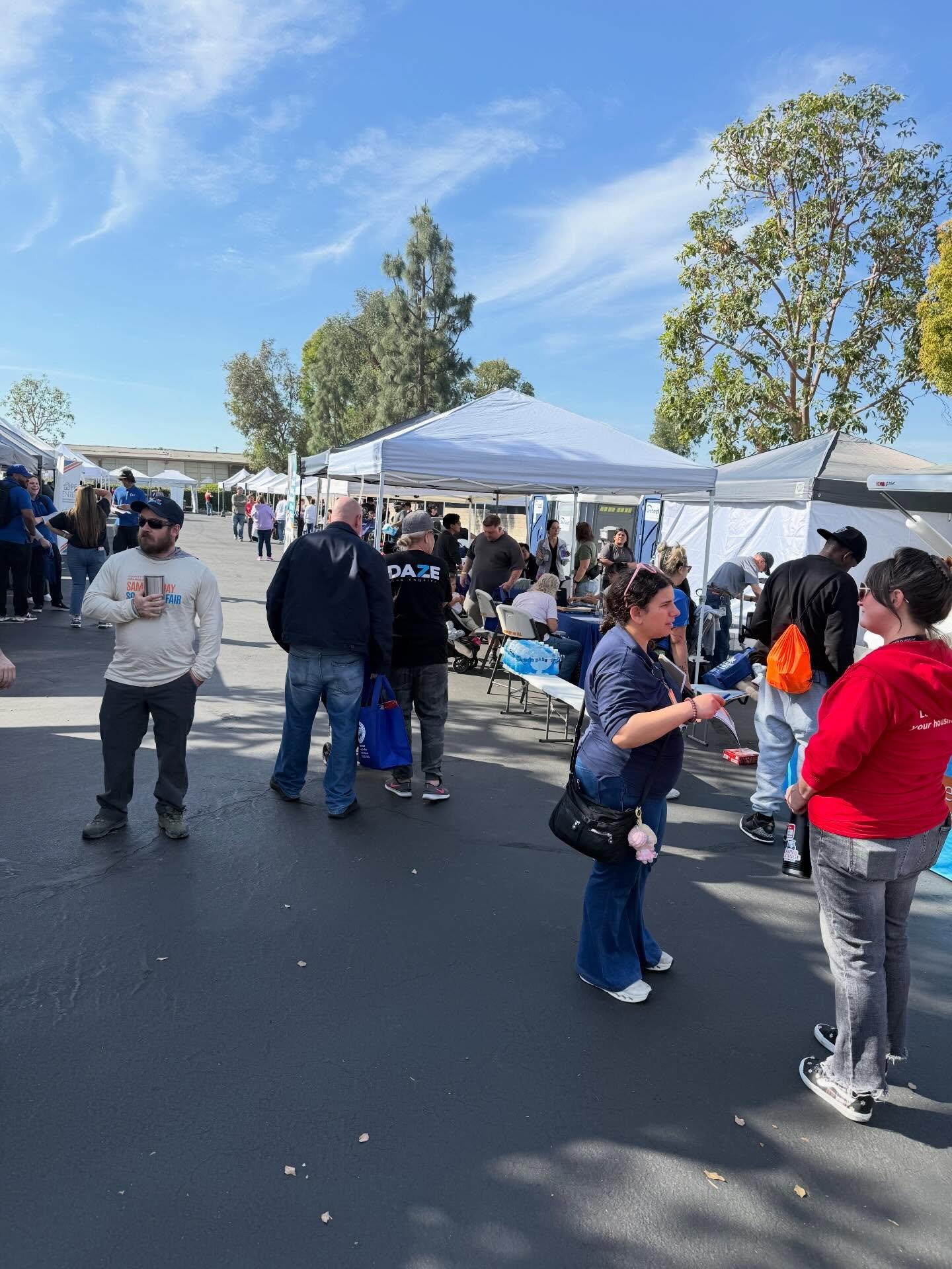 People talk and visit information booths at an outdoor fair beneath white tents.