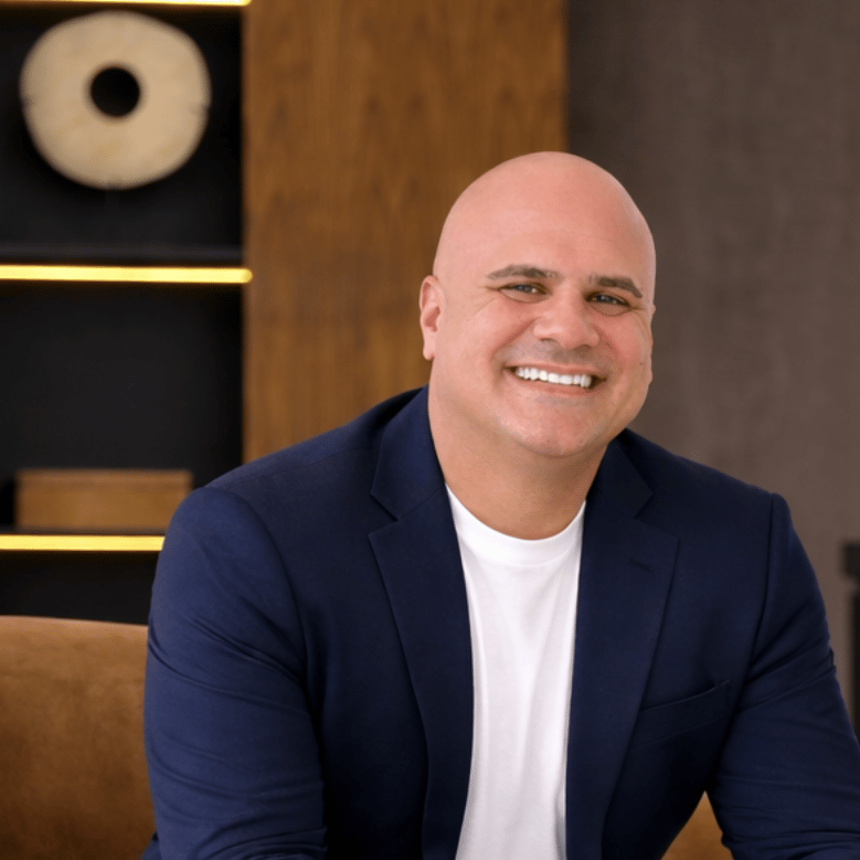 Marc Coriaty smiling in a navy blazer, seated against wood paneling.