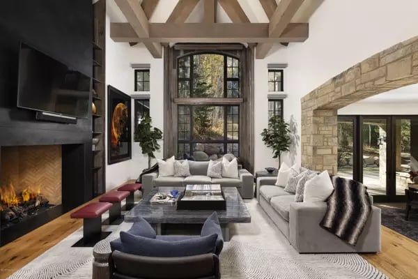 Living room with exposed ceiling beams, a tall window, and a stone fireplace beside neutral sofas.