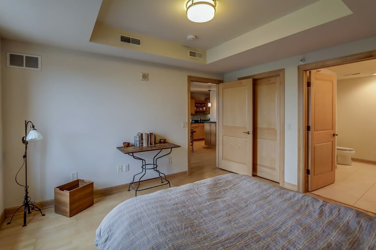 Bedroom with a bed, small book-topped table, and open doors to a kitchenette and bathroom.