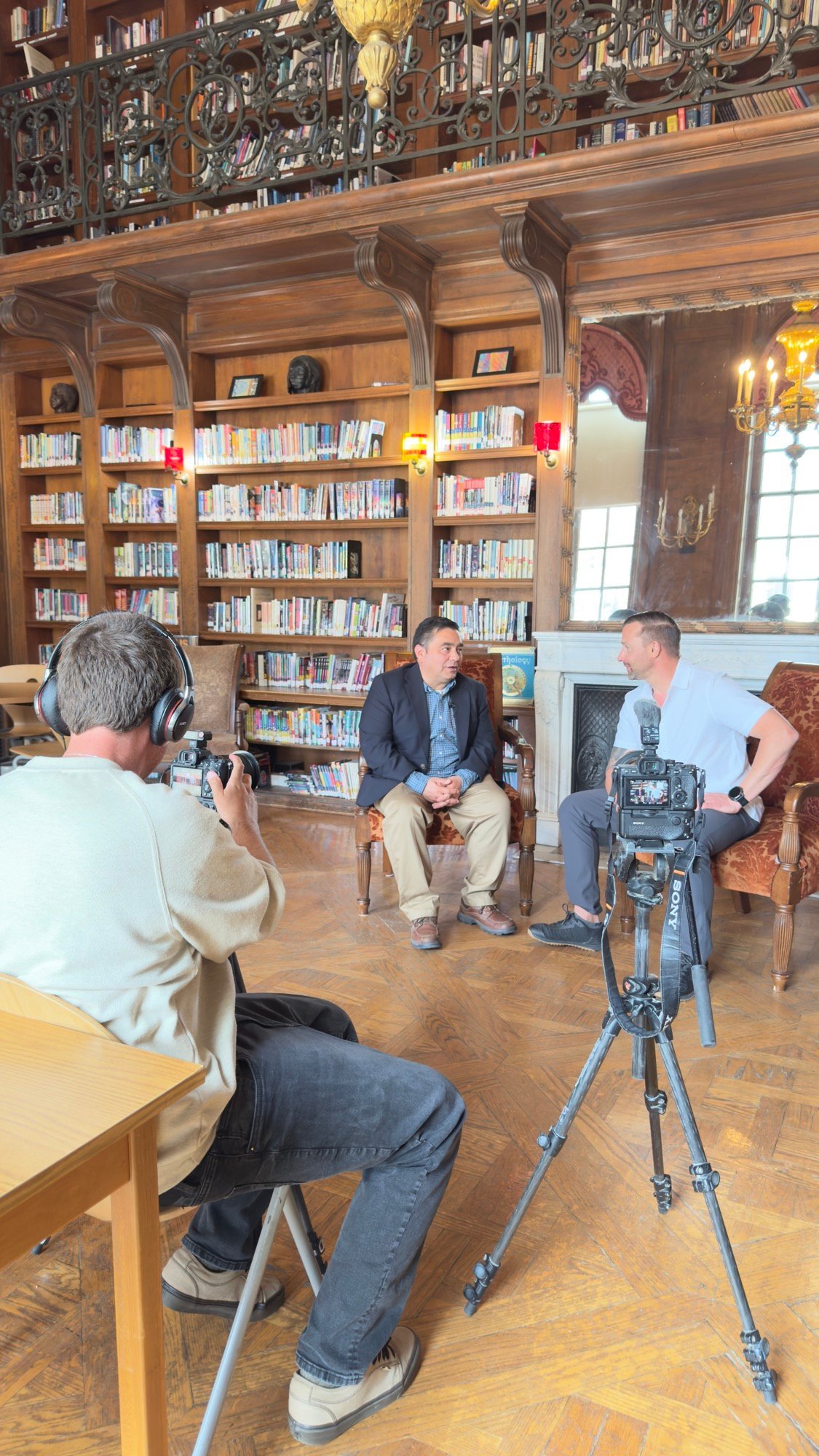 Two men talk on camera in a wood-paneled library as a cameraman films them.