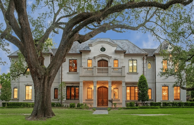 Two-story home with a central balcony, warm illuminated windows, and a large oak tree in front.