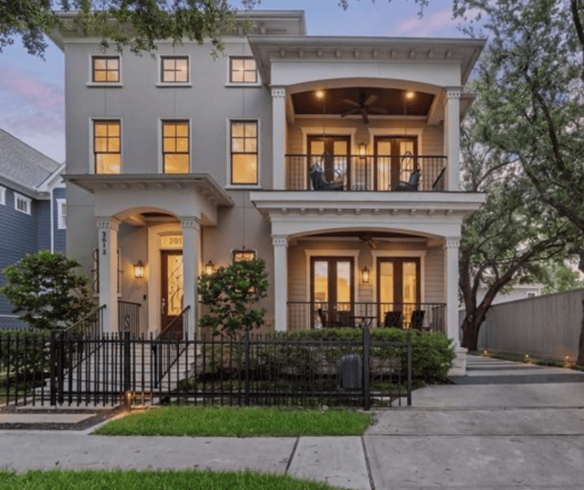 Two-story home at dusk with illuminated windows, covered balconies, and a black metal front fence.