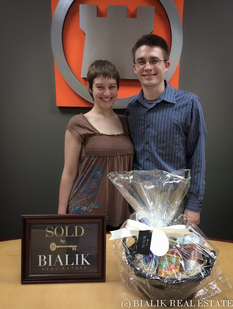 Ryan and Alycia pose with a SOLD sign and a celebratory gift basket.