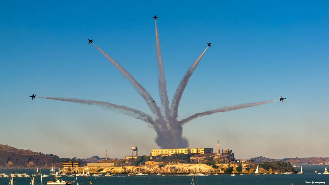 Six Blue Angels jets performing starburst break maneuver with white contrails radiating outward above Alcatraz Island, sailboats dotting San Francisco Bay waters below
