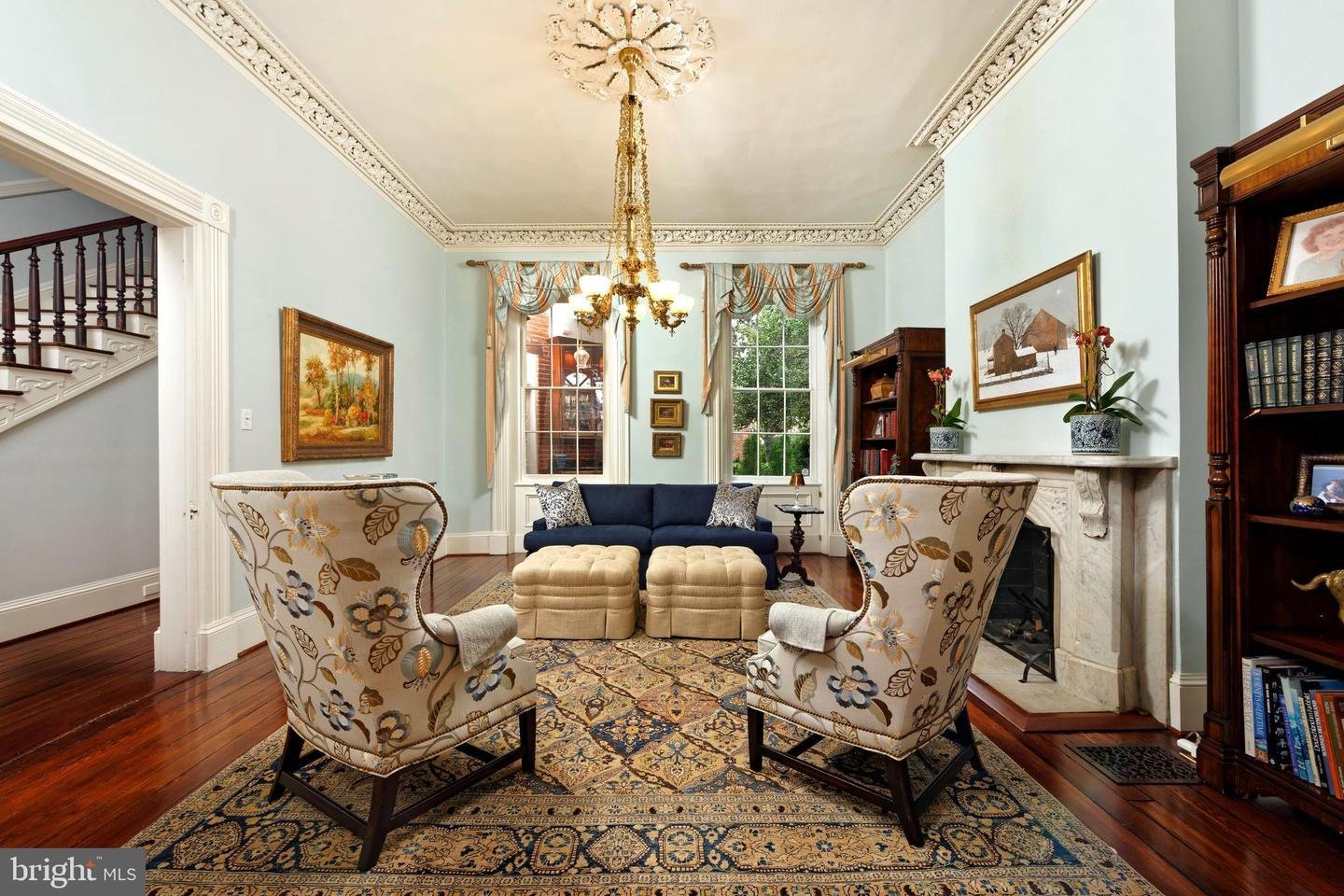 Living room with ornate ceiling molding, chandelier, marble fireplace, and floral armchairs.