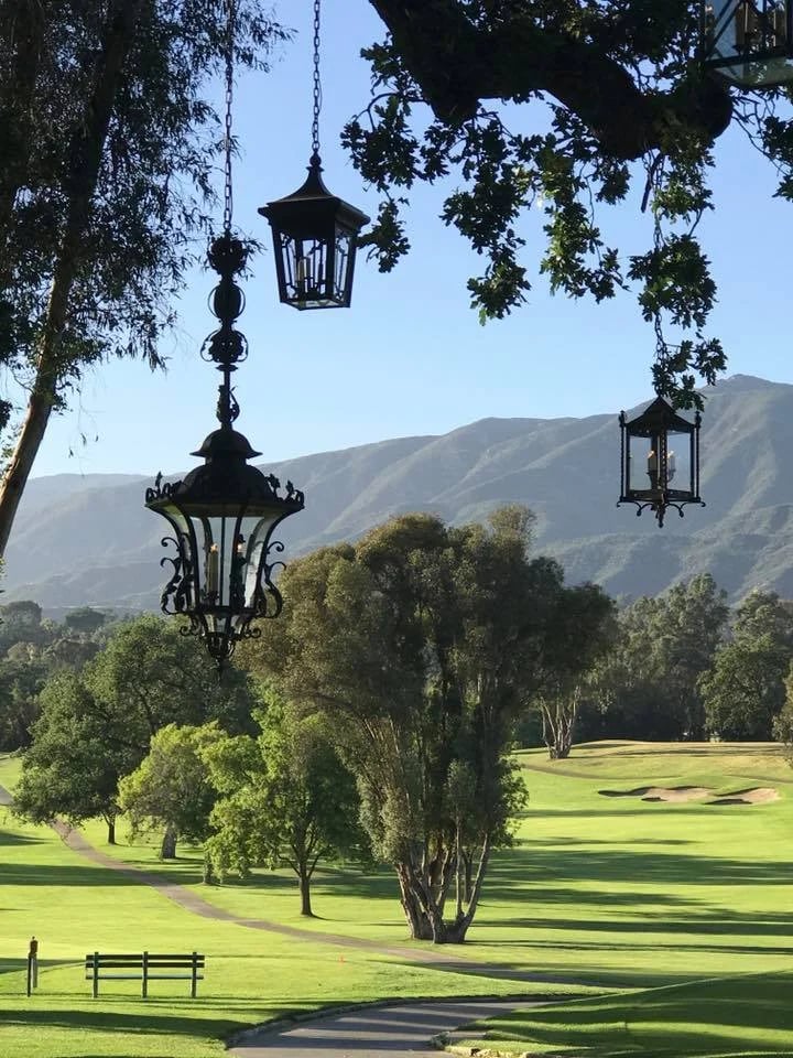 Hanging black lanterns frame a sunlit golf course with trees and mountains beyond.