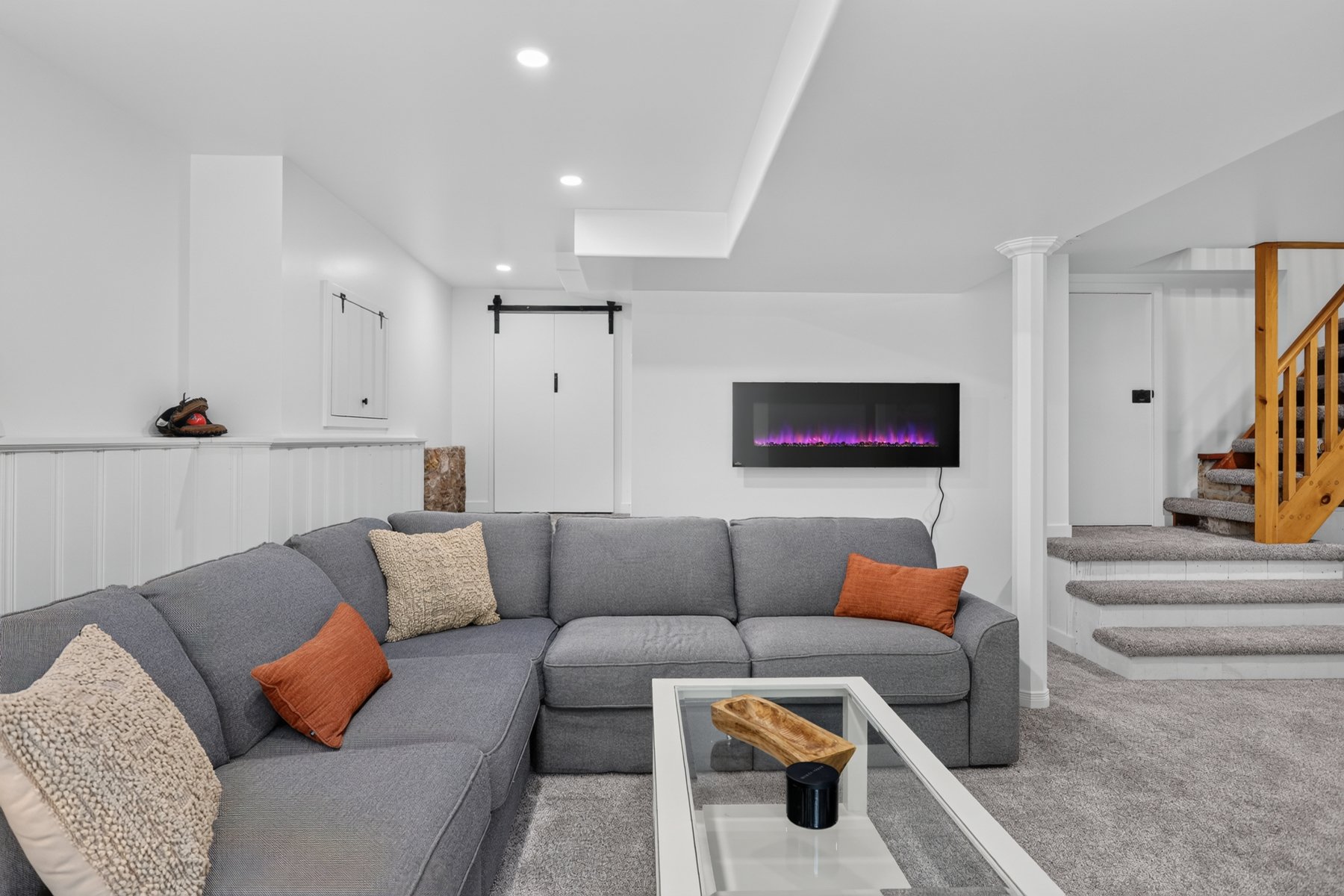 Living area with a gray sectional, purple-lit electric fireplace, and carpeted stairs.