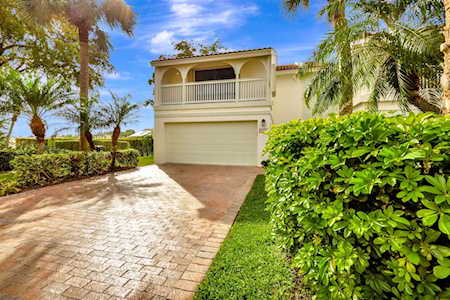 Two-story home with an upper balcony and brick-paver driveway, framed by palms and shrubs.