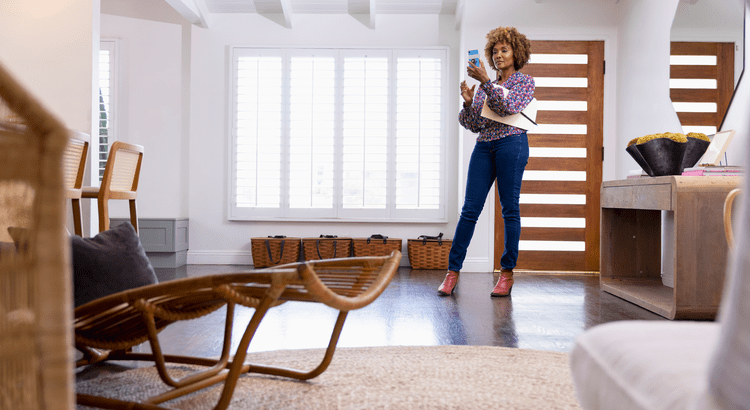 Woman in a colorful sweater and jeans stands in a bright, modern living room with wooden decor, holding a tablet and smartphone, appearing focused.