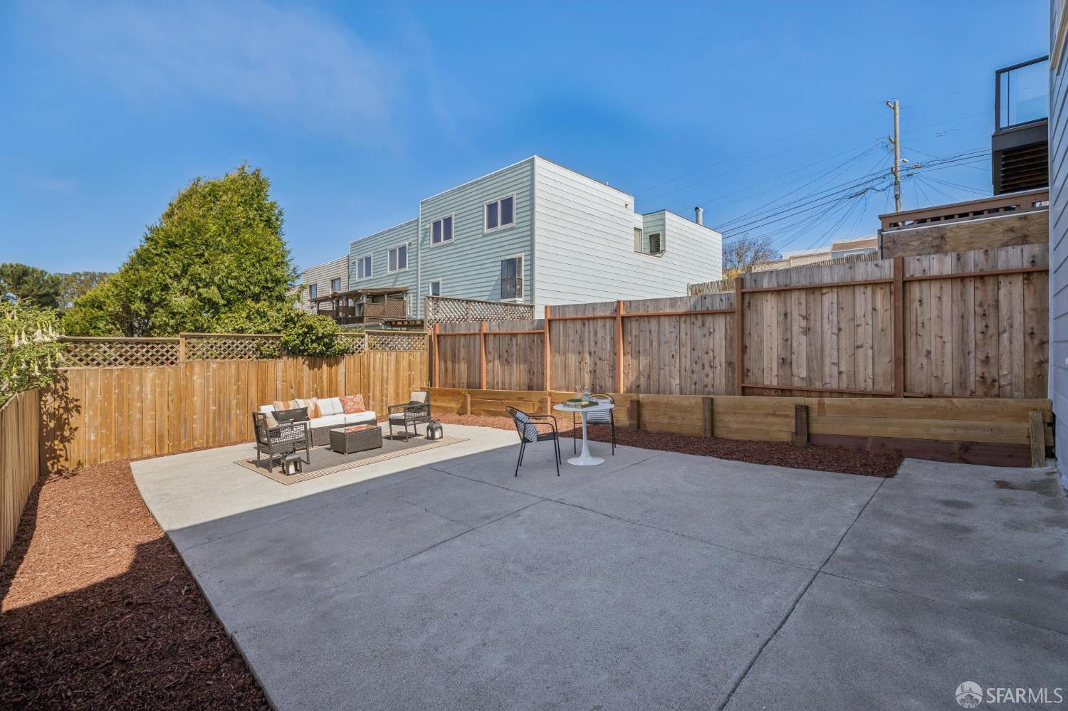 Fenced concrete patio with outdoor seating, mulched borders, and neighboring homes beyond the fence.
