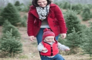 A woman in a red coat walks with a young child among evergreen trees.
