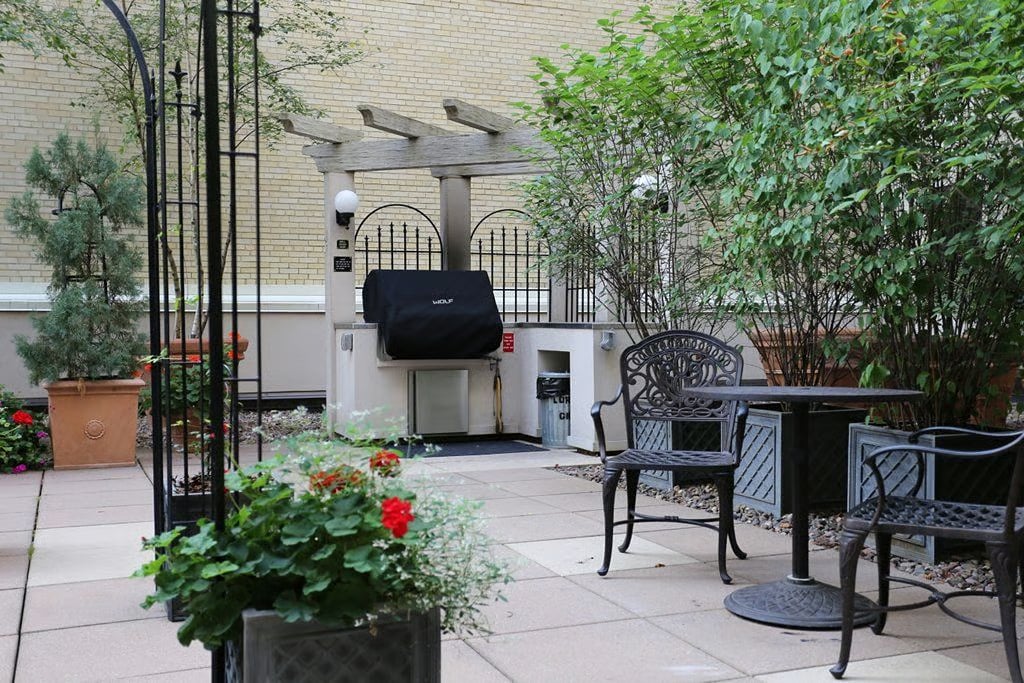 Paved courtyard patio with a built-in grill, black metal furniture, potted flowers, and shrubs.
