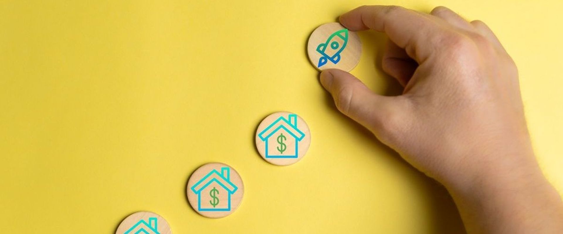 Hand places a rocket token beside wooden house-and-dollar tokens on a yellow surface.