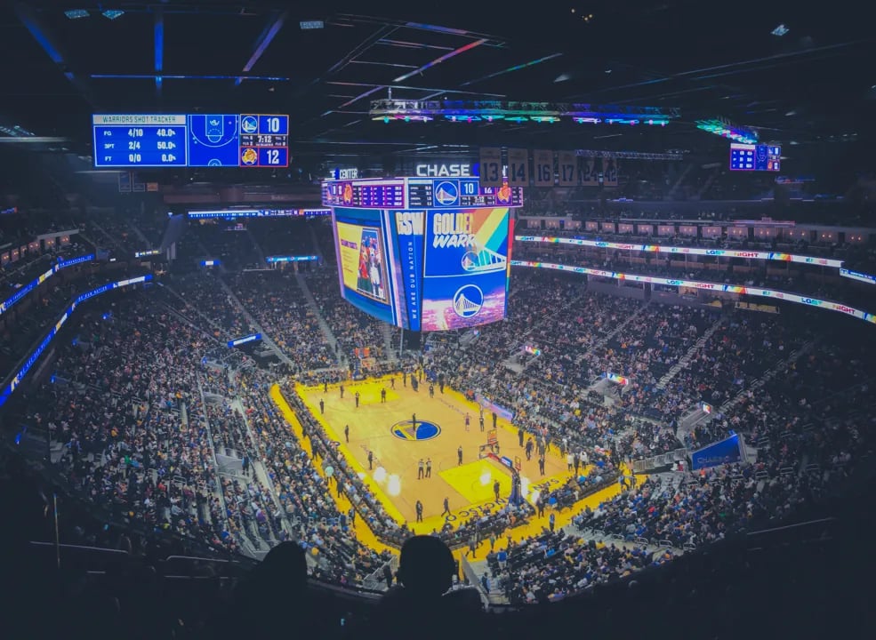 Interior shot of the Chase Center arena showing the massive LED scoreboard during a Warriors game, with the crowd in full view