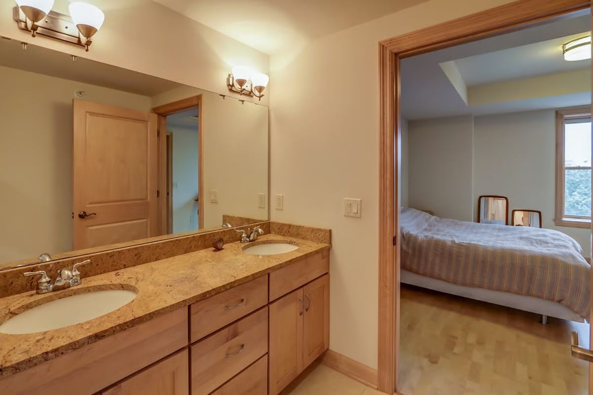Double-sink bathroom vanity with a tan stone counter opens to a bedroom with a tree-facing window.