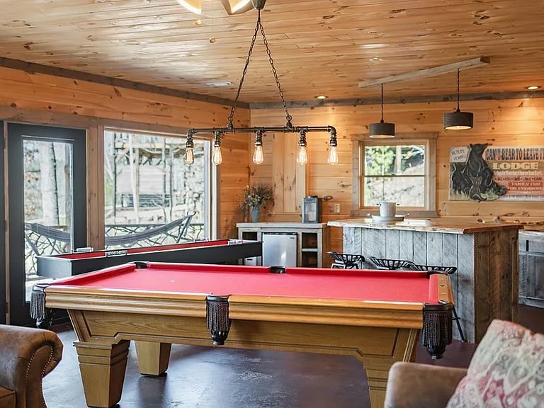 Game room with a red-felt pool table, a wood-paneled bar, and windows overlooking trees.