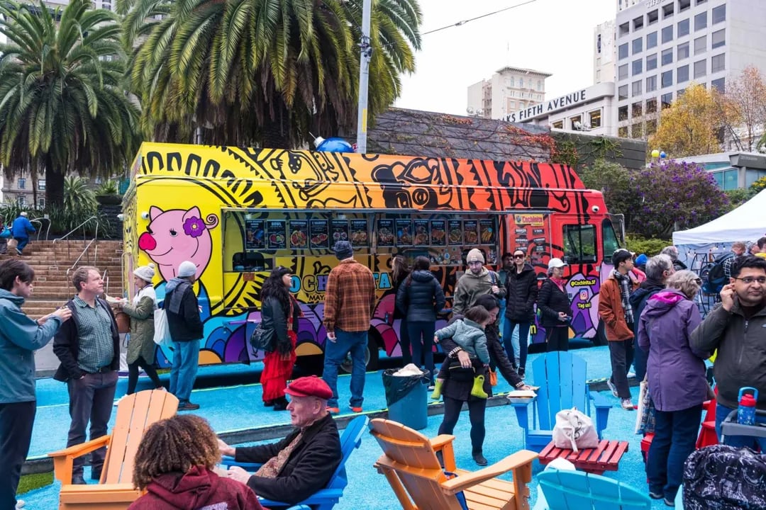  A brightly colored food truck with a graphic of a pink pig is surrounded by a crowd of people in an outdoor market setting near palm trees, with blue and orange Adirondack chairs in the foreground.