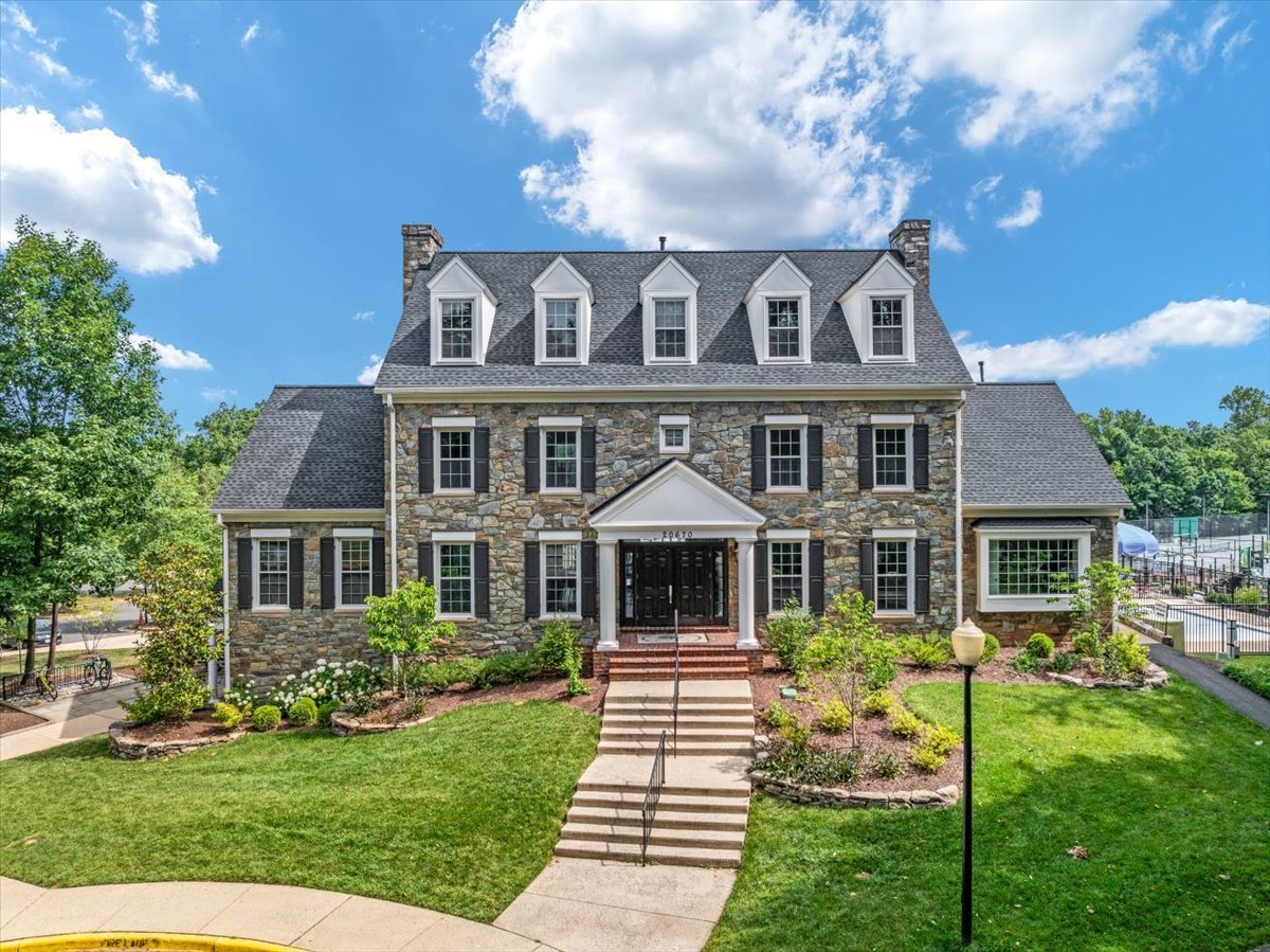 Stone house with five dormers, landscaped front lawn, and tennis court beside it.