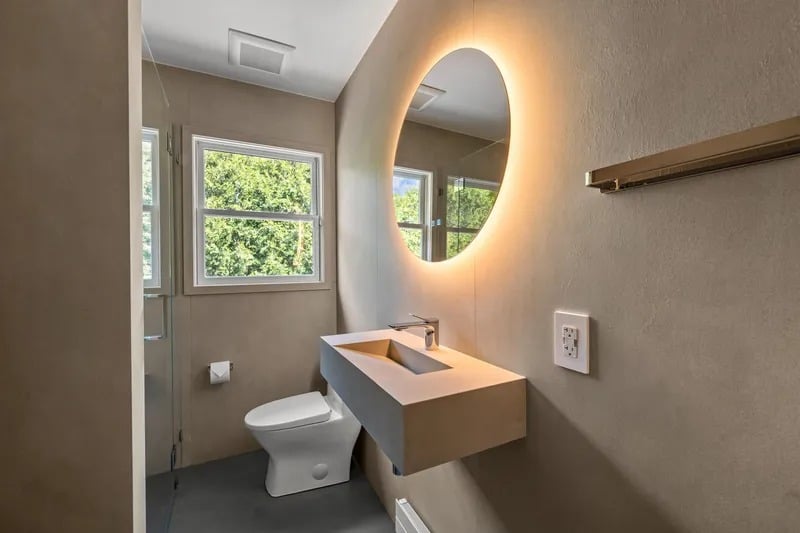 Modern bathroom with a floating vanity, backlit oval mirror, and window overlooking greenery.