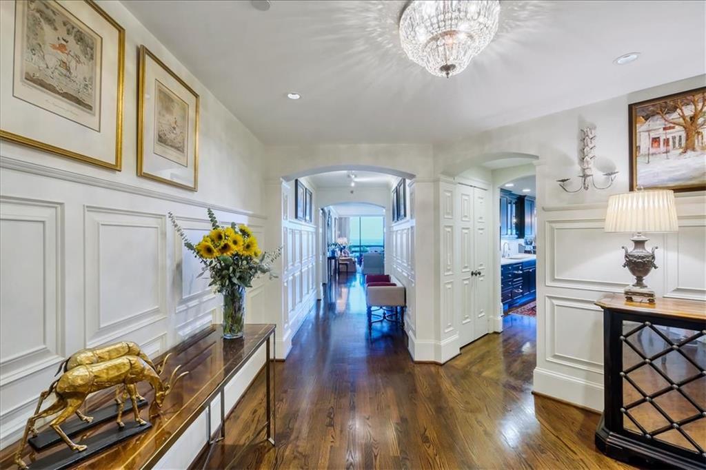 Elegant hallway with white paneled walls, polished hardwood floors, a crystal chandelier, and a distant sitting area.