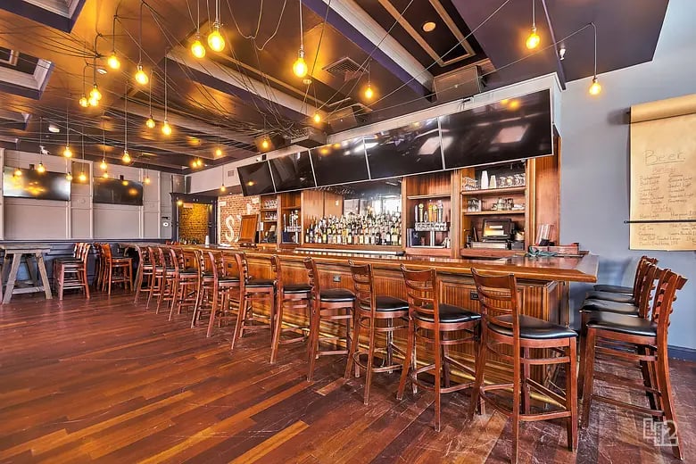 Bar interior with wooden counter, rows of stools, bottle shelves, and pendant lights.