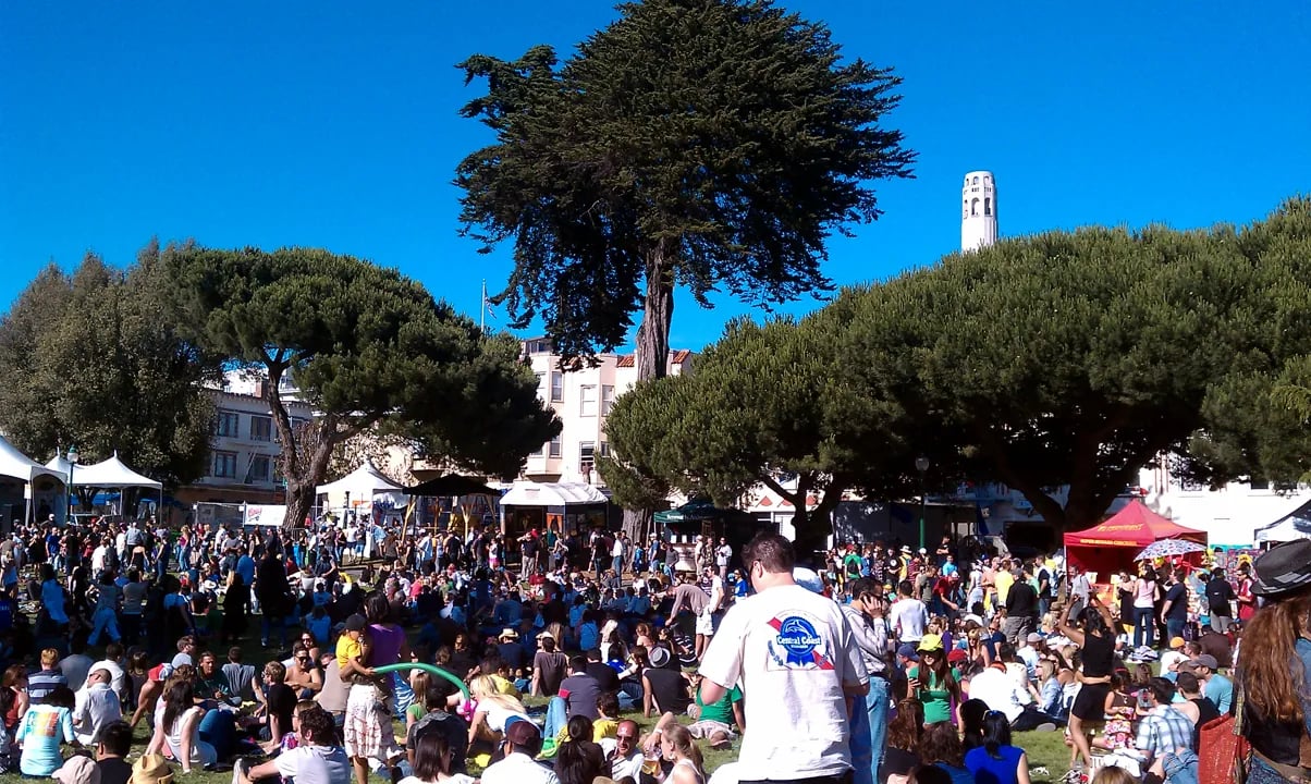 Panoramic view of Washington Square Park filled with festival tents and crowds, with Saints Peter and Paul Church and the San Francisco skyline in the background