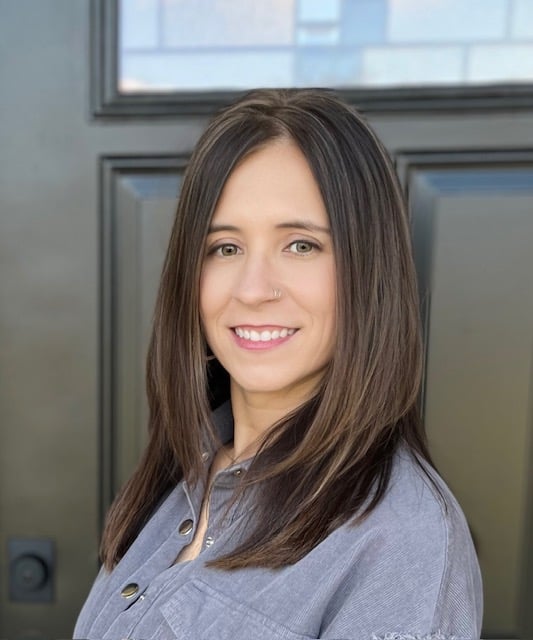 Danielle Hite smiles in a gray button-front shirt in front of a dark paneled door.