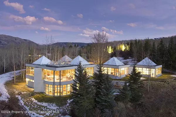 Modern multi-wing home glowing at dusk among evergreens and patches of snow.