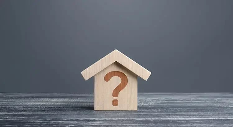 A small wooden house model with a question mark on it, placed on a wooden surface against a gray background. This image likely symbolizes questions or uncertainties about homeownership or real estate.