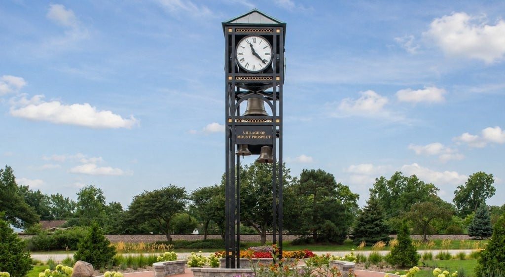 Mount Prospect clock tower with bells, surrounded by flowers and greenery.
