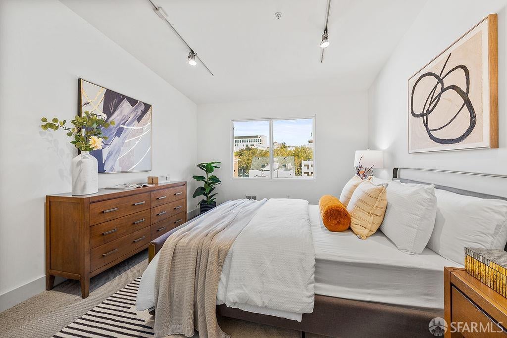 Staged bedroom with white bedding, a wooden dresser, abstract art, and a window overlooking nearby buildings.