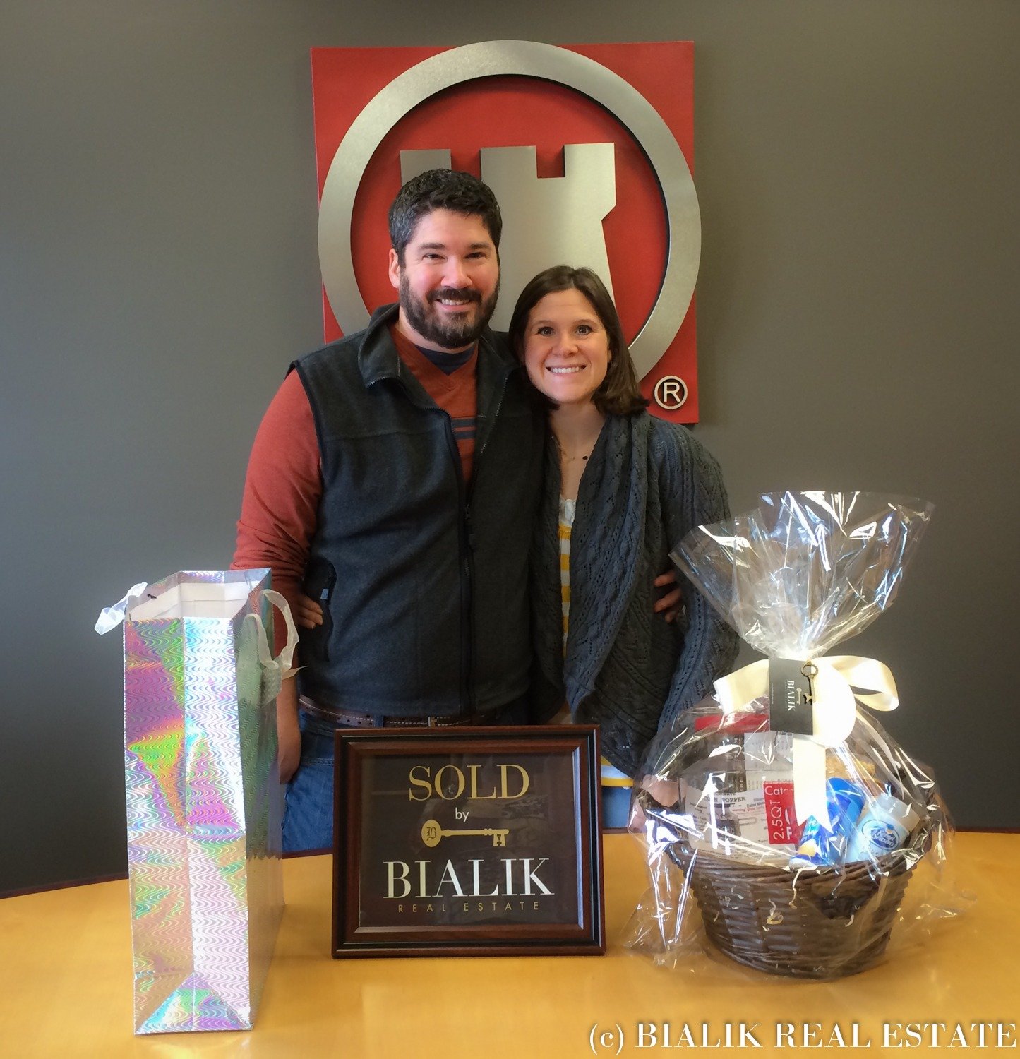 Jeff and Molly celebrate their home sale beside a SOLD sign and gift baskets.