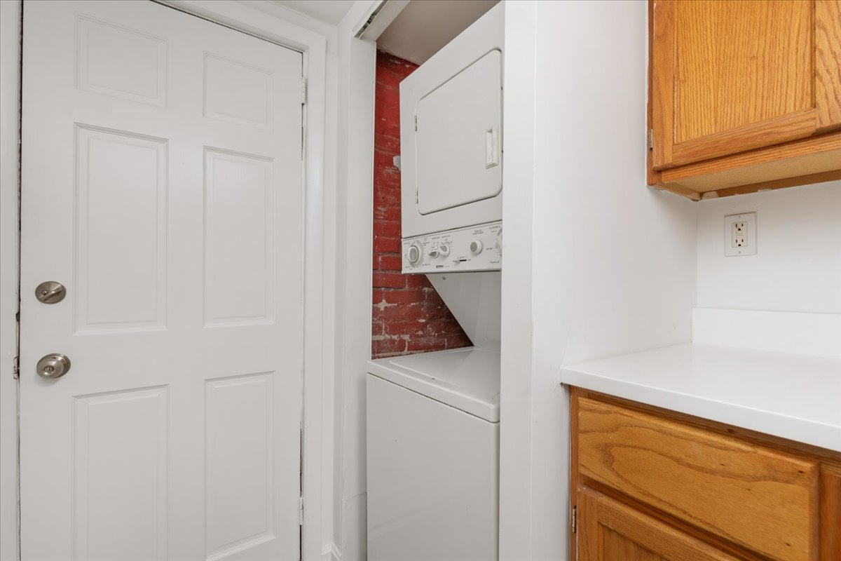 Laundry area with stacked washer and dryer, exposed brick, and wood cabinets.