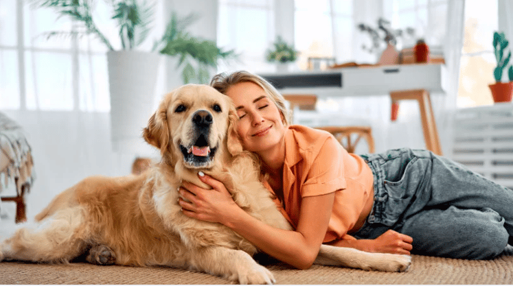 Woman cuddling a golden retriever on a rug in a bright living room.