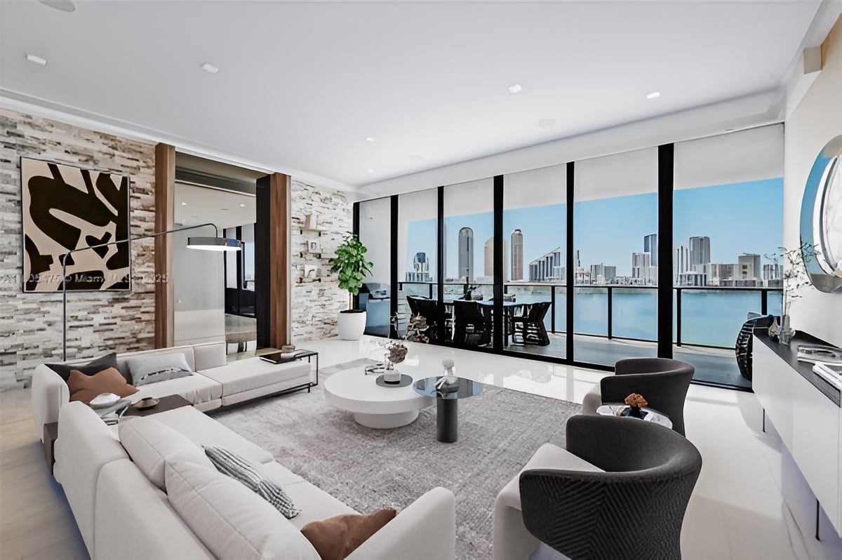 Modern living room with pale sofas and floor-to-ceiling windows overlooking water and a city skyline.