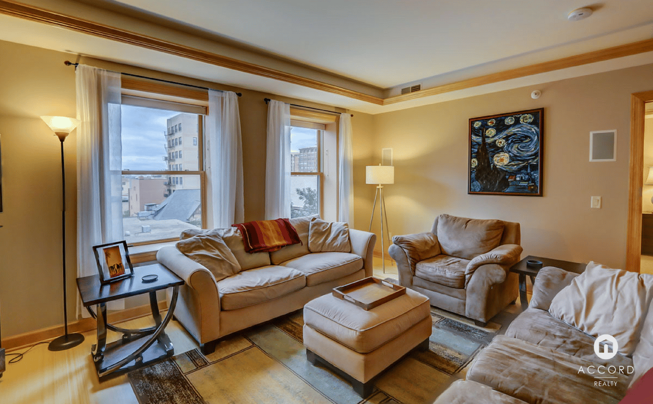 Furnished condo living room with beige seating, colorful artwork, and city buildings visible through two windows.