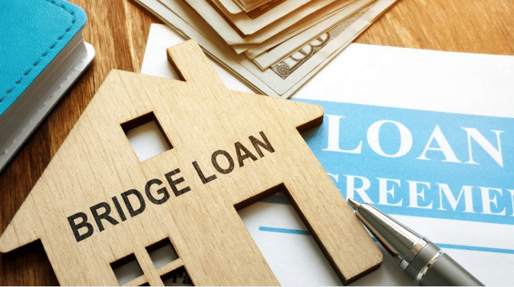 Wooden house marked “BRIDGE LOAN” on loan paperwork beside a pen and cash.