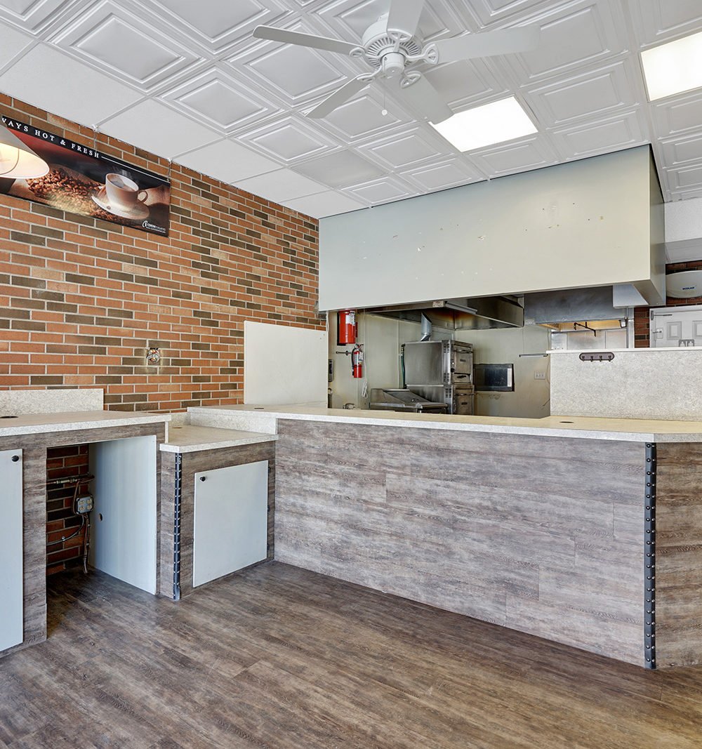 Commercial kitchen with a long wood-look service counter, exposed brick wall, and stainless-steel cooking equipment.