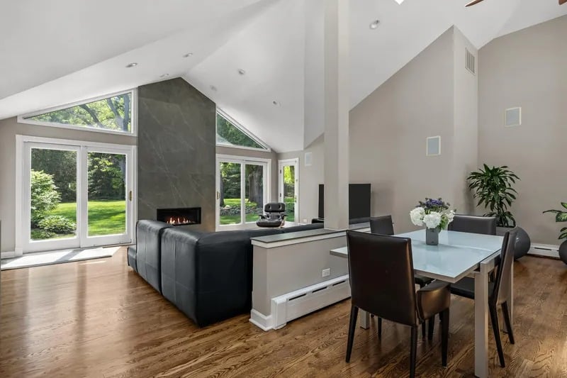 Open-plan living and dining room with vaulted ceilings, a fireplace, and large windows overlooking greenery.